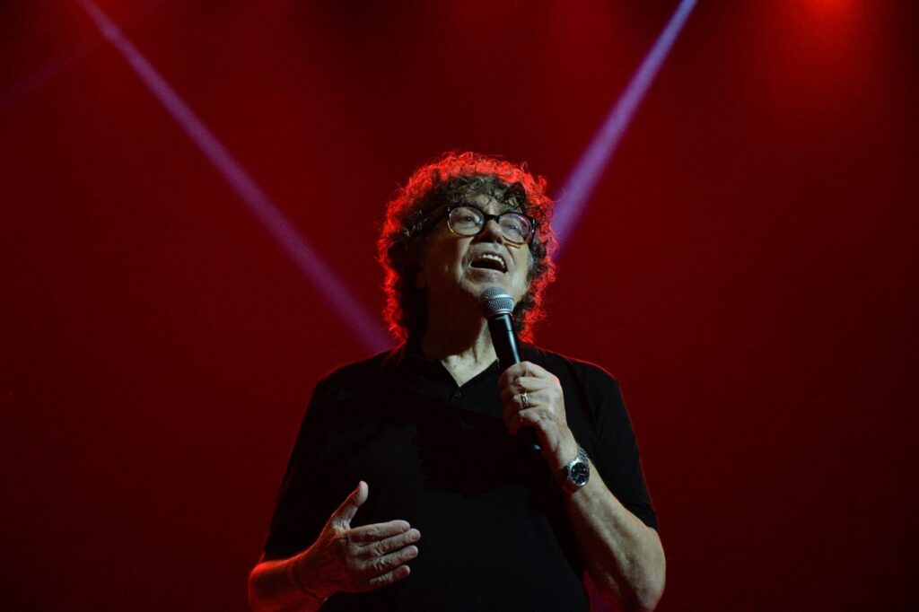 Piero canta en un escenario con un micrófono en la mano, bajo luces rojas